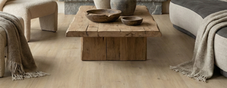Sequoia waterproof laminate flooring in a light wood finish installed in a rustic living room with a reclaimed wood coffee table, boucle armchair, and grey sofa. The durable, waterproof flooring has a realistic wood grain texture.